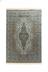 Alfombra clásica rectangular en tonos blanco suave, azul y beige con gran medallón central, arabescos florales y guardas ornamentales.