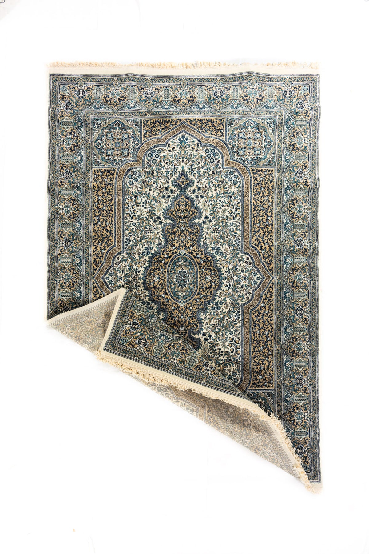 Alfombra clásica con esquina doblada que muestra el reverso, diseño ornamental en tonos blanco suave, azul y beige y flecos claros en los extremos.