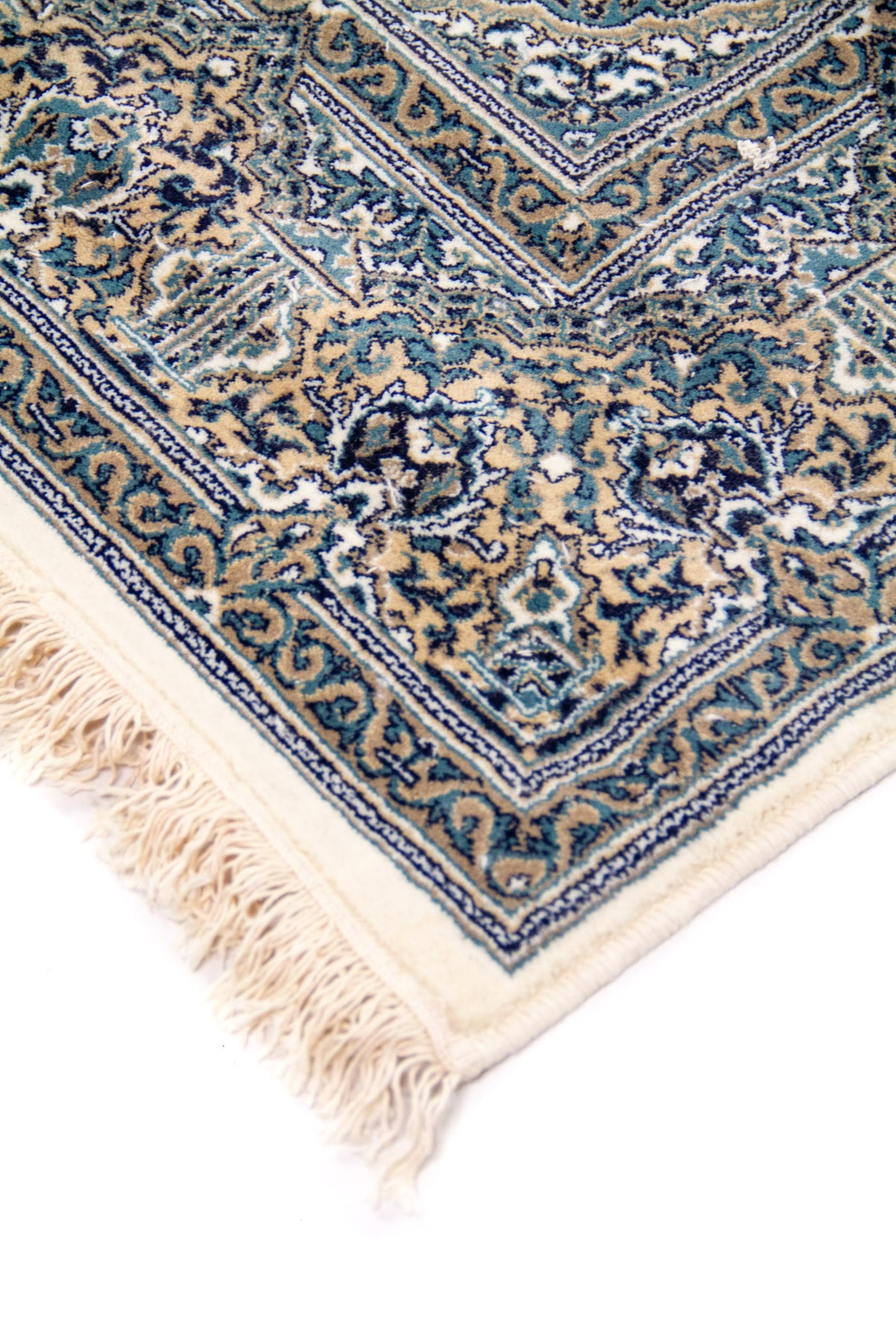 Detalle de esquina de alfombra con flecos color blanco suave, borde ornamental y delicados motivos florales en tonos azul y beige.