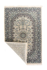 Alfombra oriental en tonos blanco suave y azul con una esquina doblada que muestra el reverso, los flecos y el detalle del tejido.