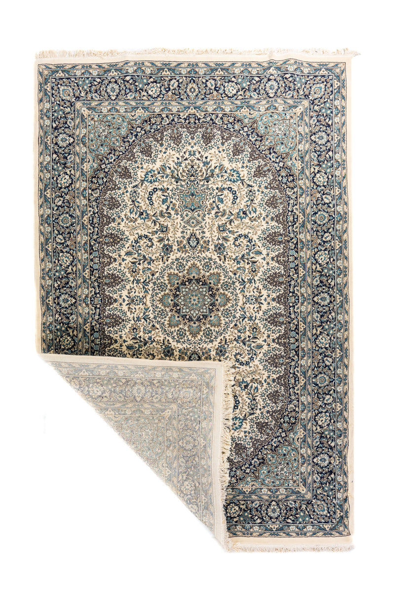 Alfombra oriental en tonos blanco suave y azul con una esquina doblada que muestra el reverso, los flecos y el detalle del tejido.