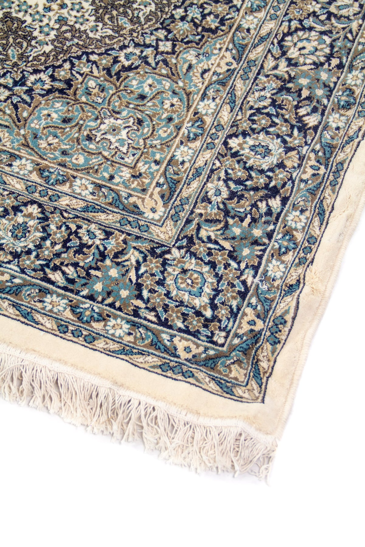 Detalle de esquina de alfombra clásica con guarda floral azul, fondo blanco suave y flecos claros en uno de sus extremos