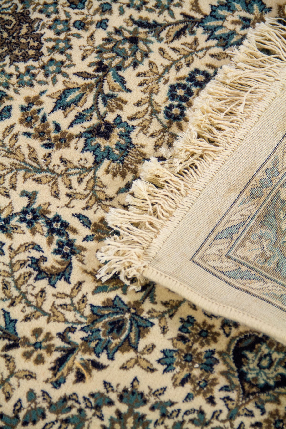 Detalle de alfombra doblada mostrando los flecos, el reverso y el diseño floral de inspiración persa en tonos azul y beige.
