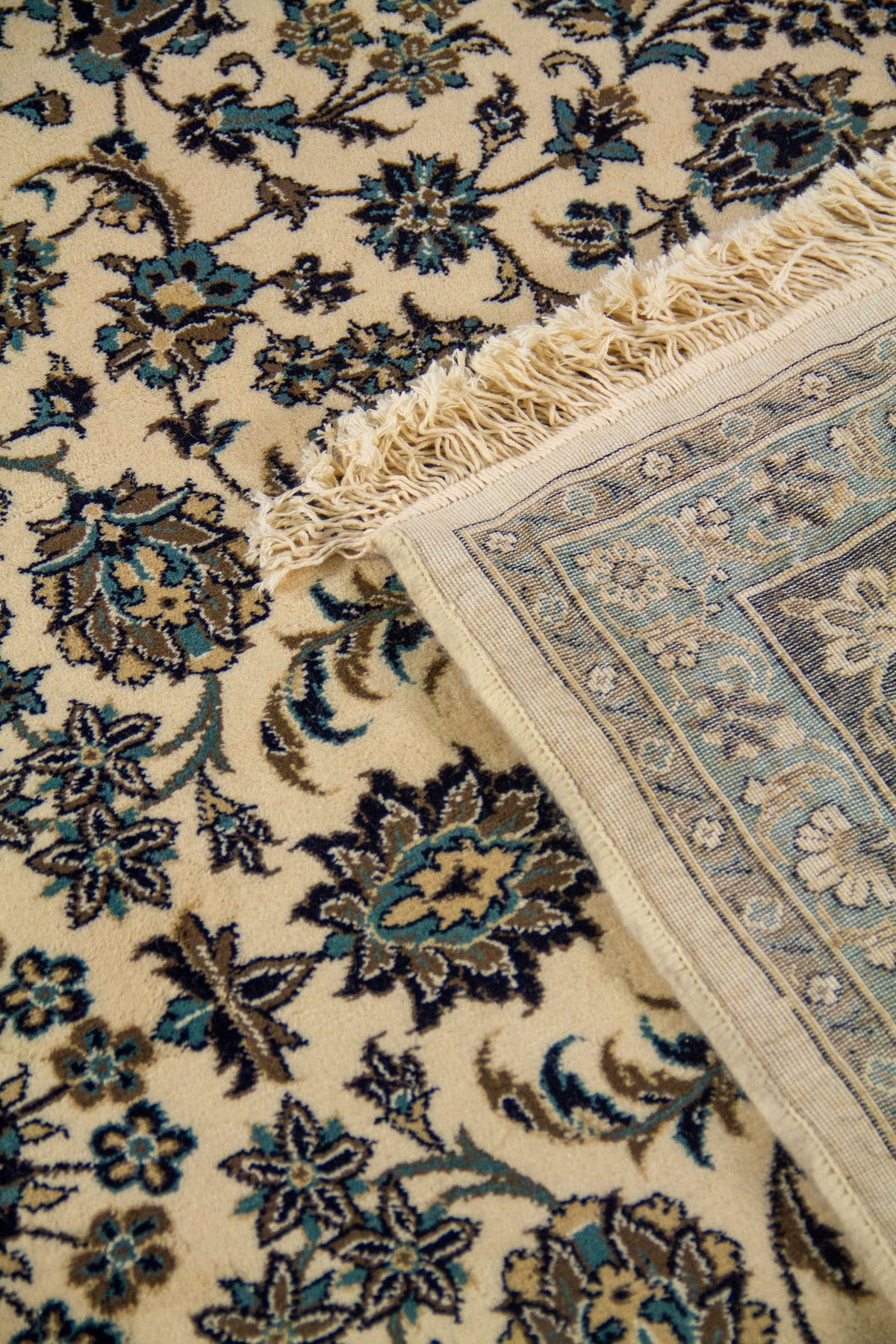 Detalle de la alfombra doblada con reverso visible, flecos claros y diseño floral fino en tonos azul oscuro, celeste y blanco suave.