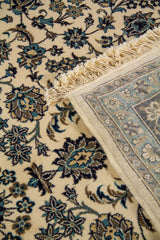 Detalle de la alfombra doblada con reverso visible, flecos claros y diseño floral fino en tonos azul oscuro, celeste y blanco suave.