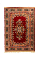 Alfombra clásica oriental en tonos rojo intenso, beige y azul con medallón central floral, guarda ornamental y flecos en los extremos.