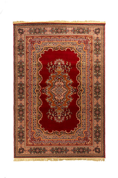 Alfombra clásica oriental en tonos rojo intenso, beige y azul con medallón central floral, guarda ornamental y flecos en los extremos.