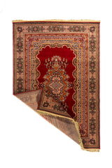 Alfombra oriental roja con una esquina doblada que deja ver el reverso, el tejido interior, la guarda decorativa y los flecos beige.