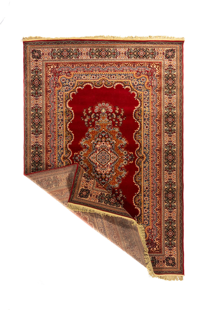 Alfombra oriental roja con una esquina doblada que deja ver el reverso, el tejido interior, la guarda decorativa y los flecos beige.