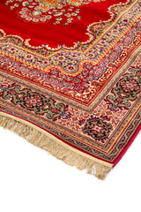 Detalle en perspectiva de alfombra clásica roja con diseño floral, borde ornamental en tonos beige y azul, y flecos claros en el extremo.