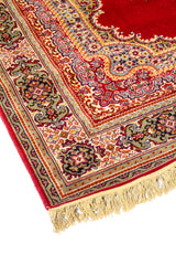 Acercamiento lateral de alfombra oriental roja con motivos florales curvos, cenefa decorativa y terminación con flecos beige.