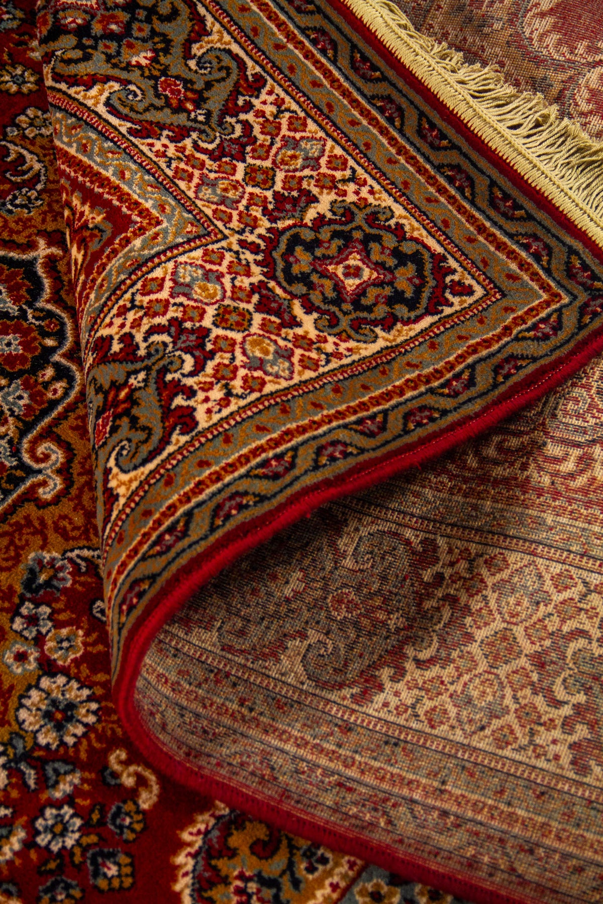 Detalle de alfombra oriental parcialmente doblada mostrando el reverso, la textura del tejido y la riqueza ornamental del borde.