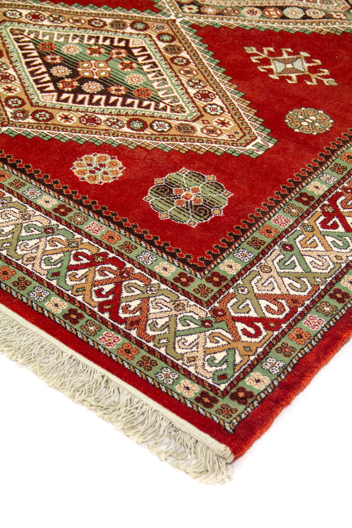 Detalle de esquina de alfombra rojo con medallón geométrico, borde ornamental en verde y beige, y flecos claros