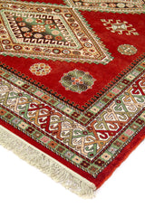 Detalle de esquina de alfombra rojo con medallón geométrico, borde ornamental en verde y beige, y flecos claros