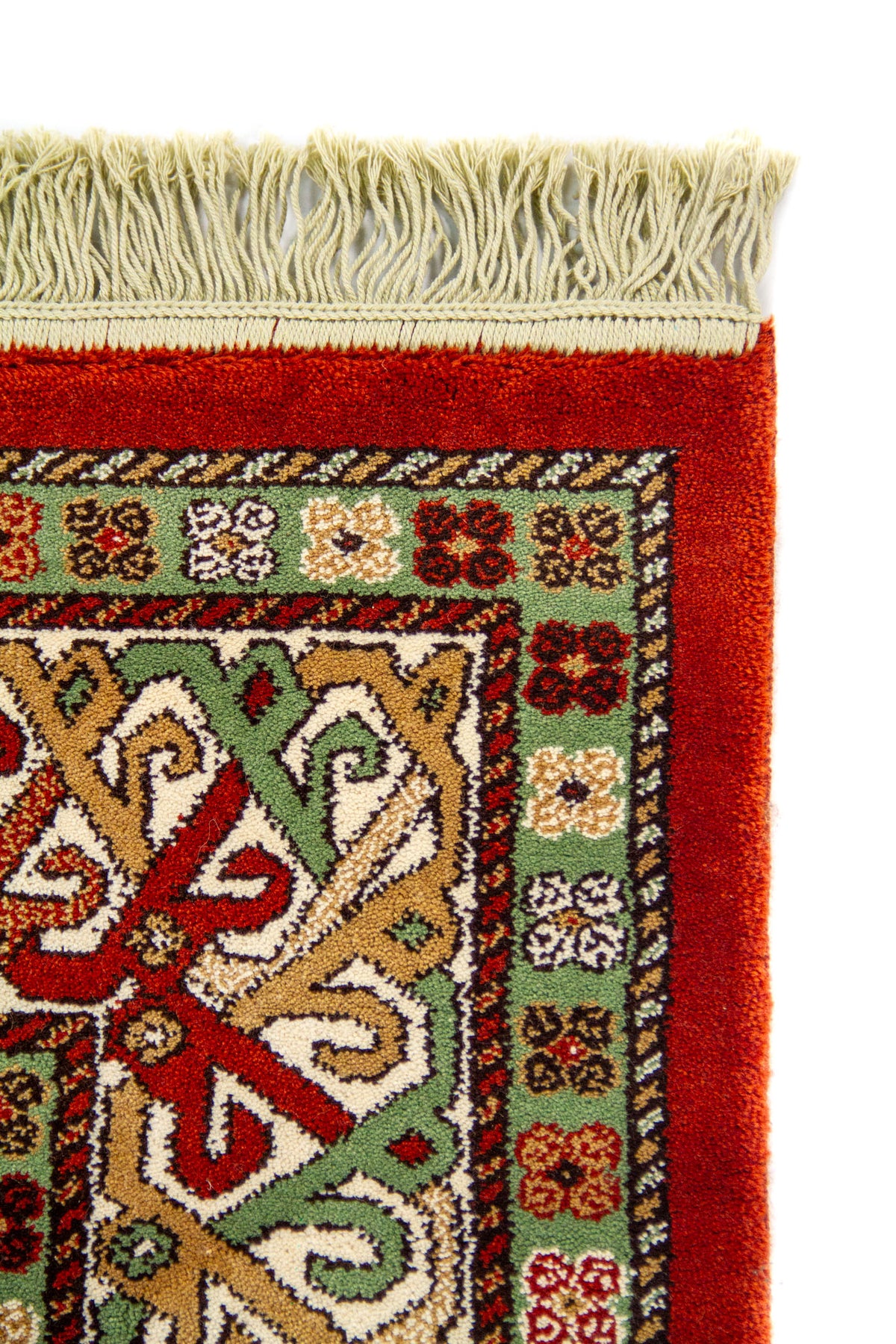 Detalle de esquina superior de alfombra clásica con flecos beige y guarda geométrica en tonos verde, blanco suave, rojo y marrón