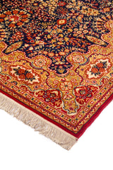 Detalle en perspectiva de alfombra clásica con fondo azul oscuro, motivos florales en tonos dorados y rojos, y flecos claros en el borde.