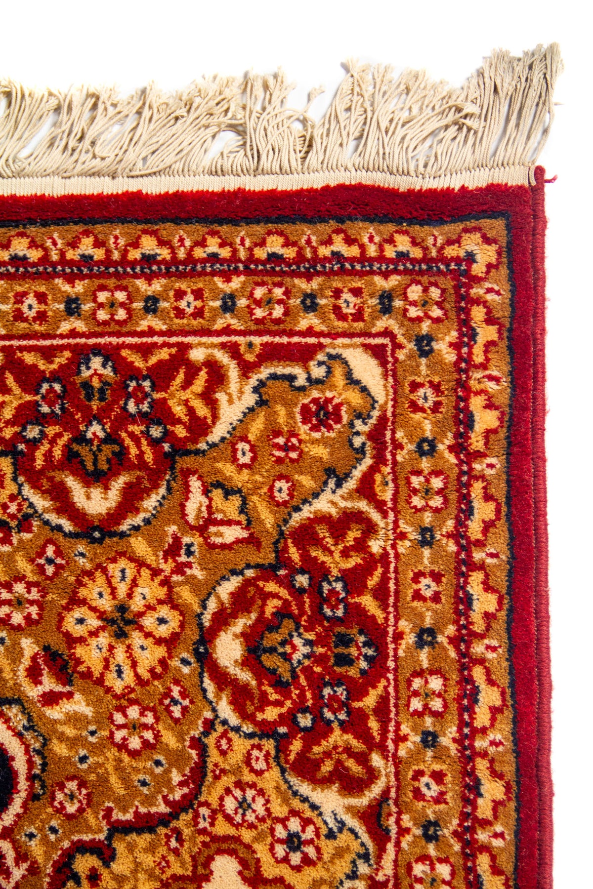Primer plano de la esquina de una alfombra clásica con borde rojo, guarda floral en tonos dorados y beige, y flecos claros.