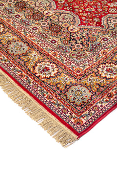 Detalle en perspectiva de alfombra roja clásica con motivos florales, cenefa ornamental en tonos beige, azul y dorado, y flecos en el extremo.