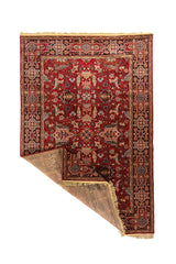 Alfombra oriental roja con una esquina doblada que muestra el reverso, la estructura del tejido, la guarda decorativa y los flecos beige.