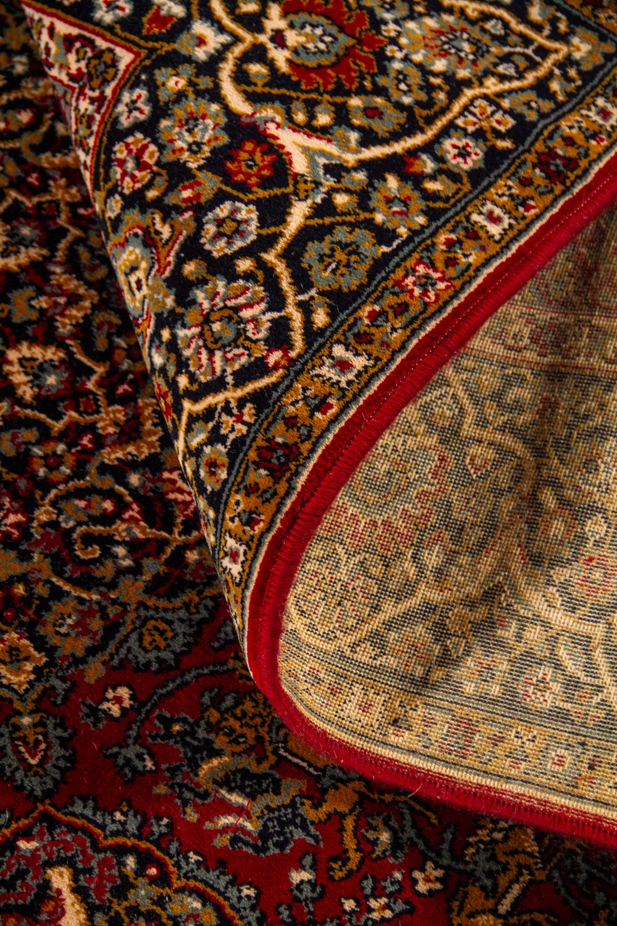 Detalle de alfombra doblada mostrando el reverso del tejido, borde rojo y patrones florales tradicionales.