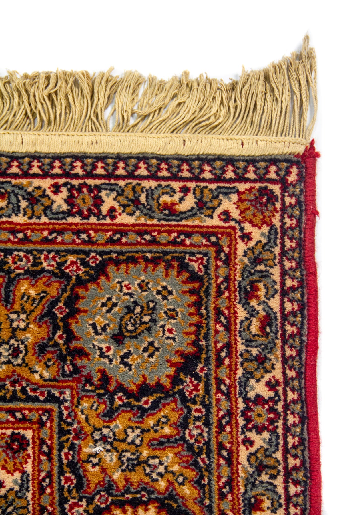 Detalle de esquina superior de alfombra oriental con flecos beige y guarda decorativa en tonos rojo, azul y beige.