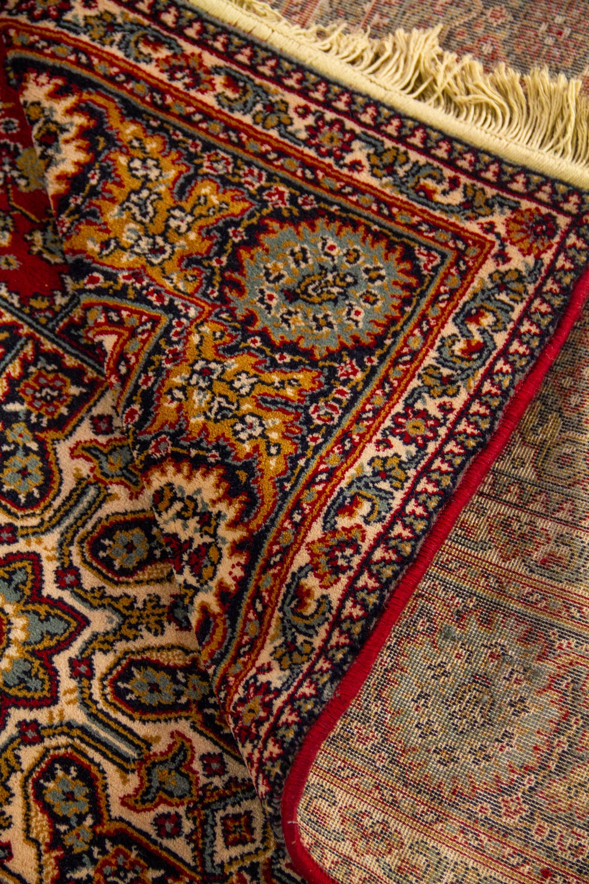 Detalle de alfombra doblada mostrando textura, reverso del tejido y diseño floral ornamental en tonos rojo, beige y azul