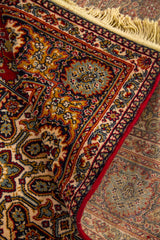 Detalle de alfombra doblada mostrando textura, reverso del tejido y diseño floral ornamental en tonos rojo, beige y azul