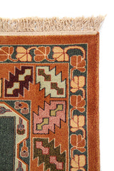 Detalle lateral de alfombra clásica con guarda geométrica en tonos anaranjado oscuro, verde, beige y marrón, y terminación con flecos