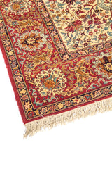 Primer plano de borde de alfombra oriental con fondo rojo, motivos florales ornamentales y acabado con flecos en tono beige.