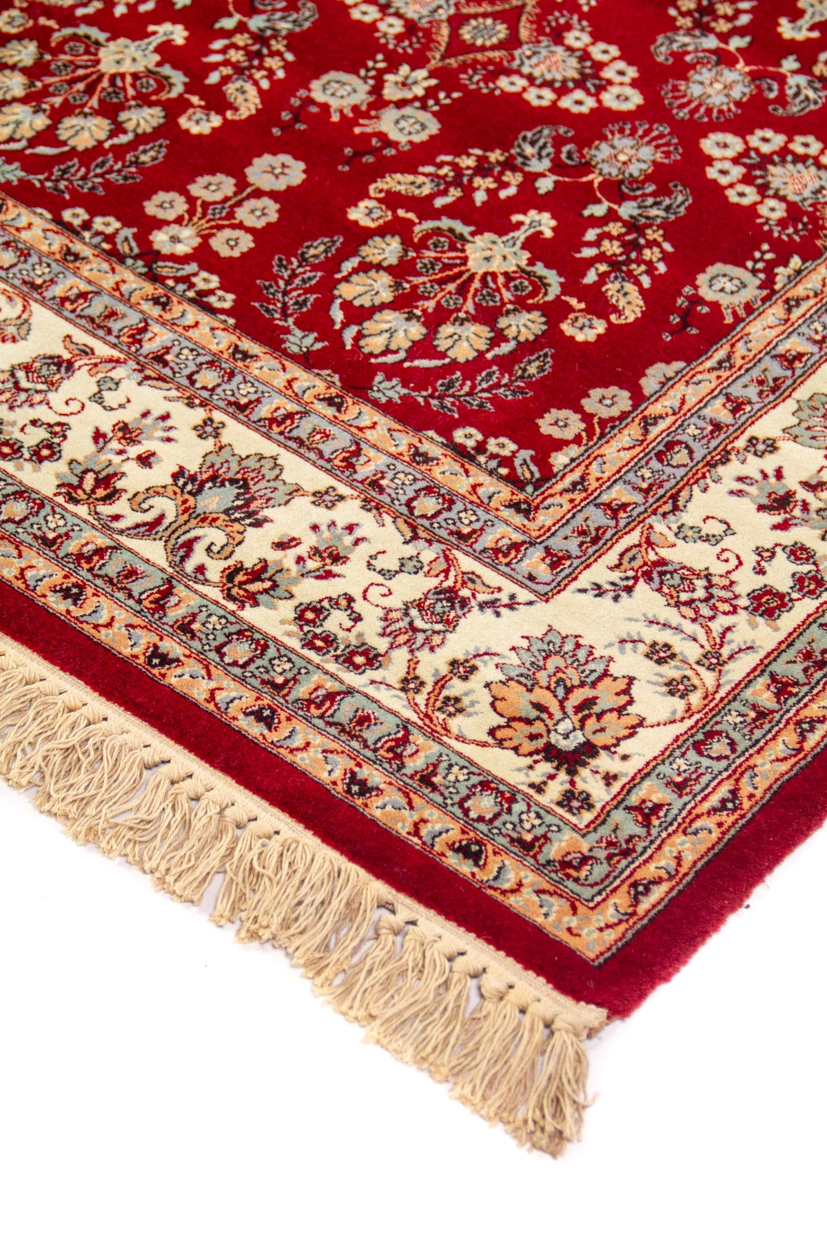 Detalle de esquina de alfombra roja con flecos beige, guarda floral ornamental y delicados motivos en tonos blanco suave, azul y dorado.