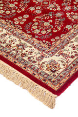 Detalle de esquina de alfombra roja con flecos beige, guarda floral ornamental y delicados motivos en tonos blanco suave, azul y dorado.