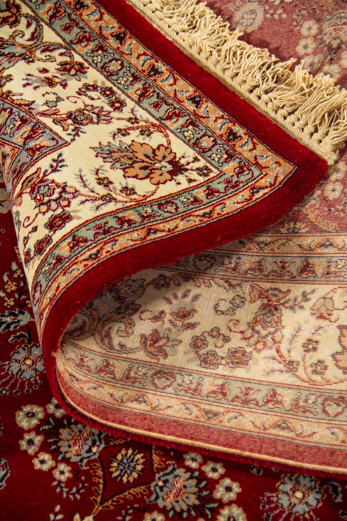 Alfombra parcialmente enrollada que muestra la textura, el reverso tejido y el diseño floral tradicional en rojo, beige y tonos suaves.