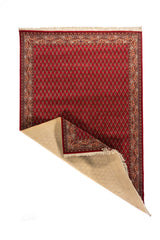 Alfombra clásica roja con una esquina doblada que deja ver el reverso, diseño repetitivo de pequeños motivos ornamentales y borde floral tradicional en tonos blanco suave y azul.