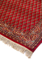 Detalle de esquina de alfombra roja con flecos beige, patrón repetitivo minucioso en el campo central y guarda ornamental con arabescos en rojo, azul marino y blanco suave.