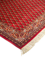 Primer plano de borde de alfombra tradicional roja con diseño repetitivo de pequeños motivos y guarda decorativa elaborada en tonos blanco suave, azul oscuro y rojo.