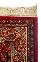 Acercamiento del extremo superior de la alfombra con flecos, mostrando el marco ornamental y la riqueza del tejido en tonos rojos, beige y azul