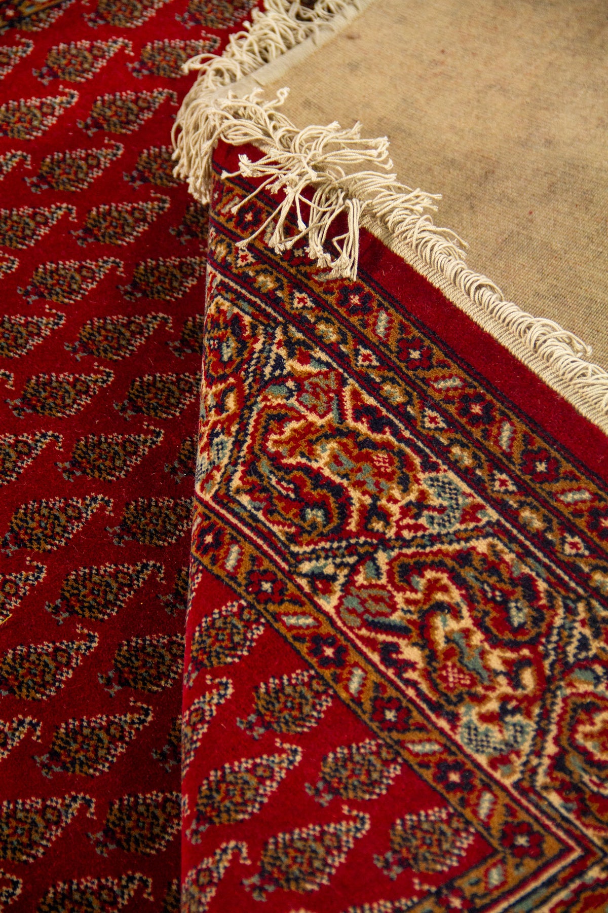 Detalle de alfombra doblada mostrando el reverso, los flecos y la transición entre el campo rojo con motivos repetitivos y la guarda clásica decorativa.