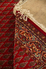 Detalle de alfombra doblada mostrando el reverso, los flecos y la transición entre el campo rojo con motivos repetitivos y la guarda clásica decorativa.