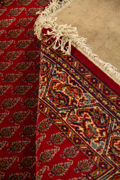 Detalle de alfombra doblada mostrando el reverso, los flecos y la transición entre el campo rojo con motivos repetitivos y la guarda clásica decorativa.