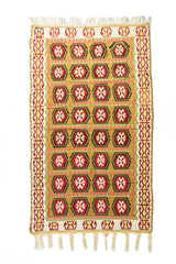 Alfombra rectangular artesanal en tonos beige, dorado, rojo y verde, con diseño geométrico repetitivo y flecos en ambos extremos.