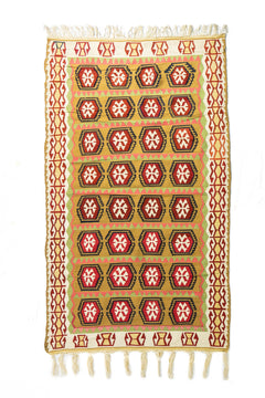 Alfombra rectangular artesanal en tonos beige, dorado, rojo y verde, con diseño geométrico repetitivo y flecos en ambos extremos.