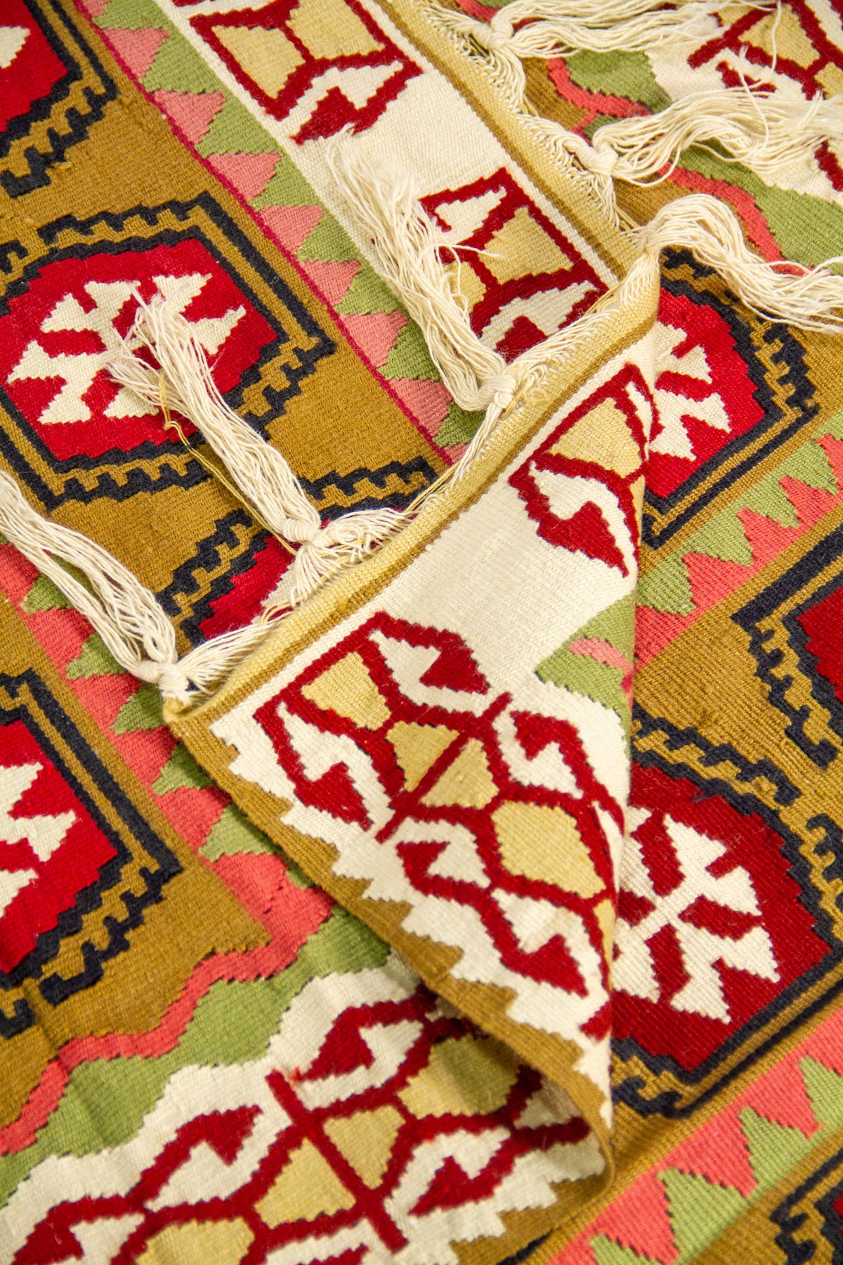 Detalle de alfombra doblada que muestra el reverso, los flecos y los patrones geométricos en tonos beige, rojo, dorado, rosa y verde.