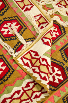 Detalle de alfombra doblada que muestra el reverso, los flecos y los patrones geométricos en tonos beige, rojo, dorado, rosa y verde.