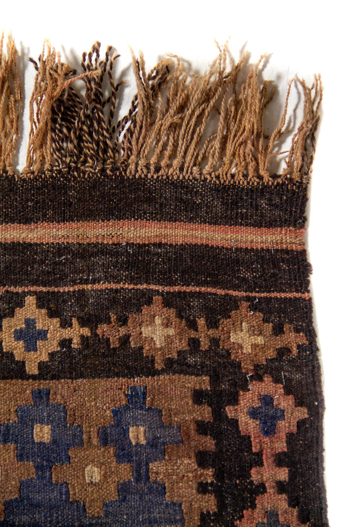 Detalle superior de alfombra tejida con flecos en tonos naturales y franjas oscuras, que resalta la textura del tejido artesanal.