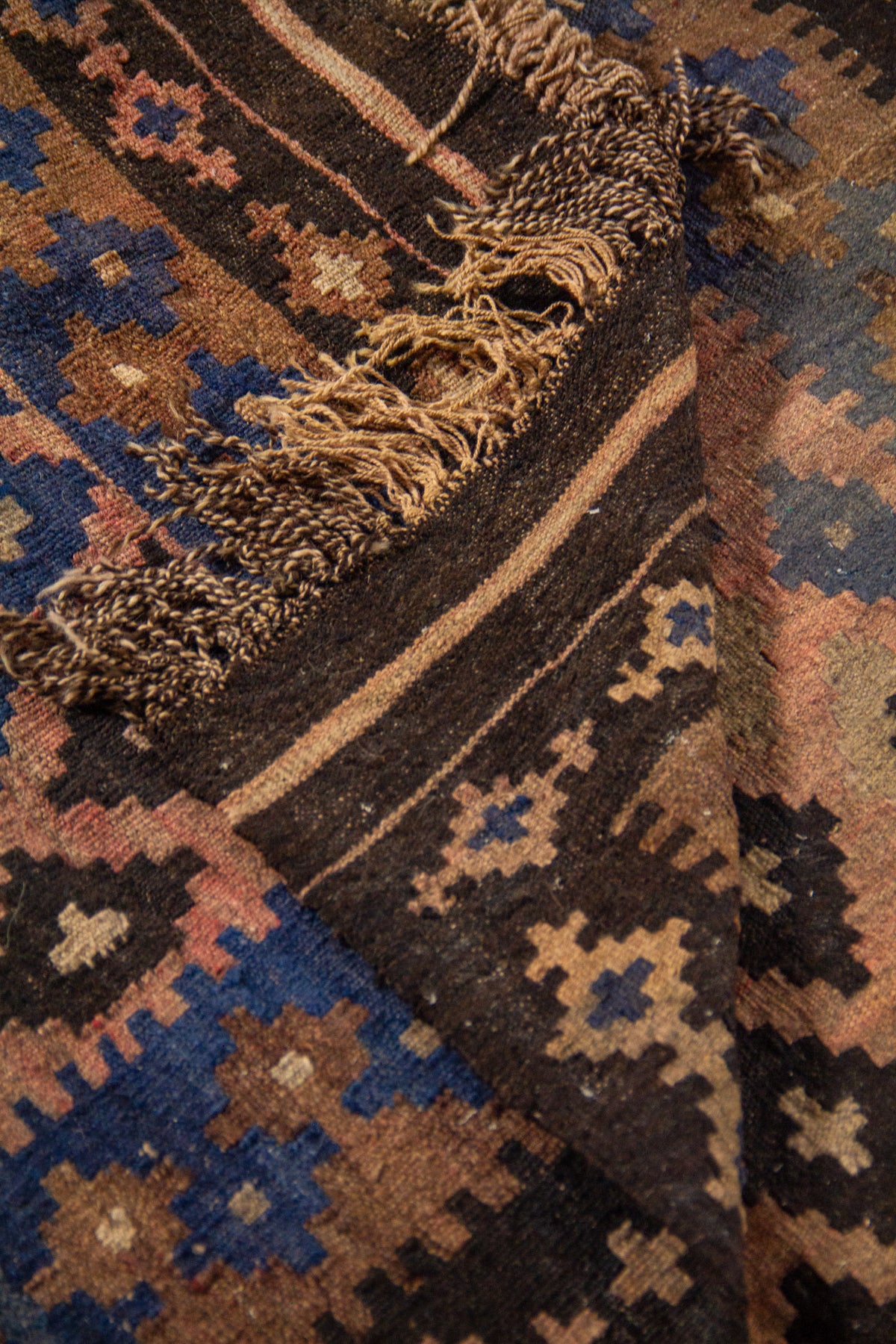 Detalle de alfombra doblada que muestra el reverso, los flecos y los patrones geométricos en tonos azul, marrón, rosa cálido y negro.