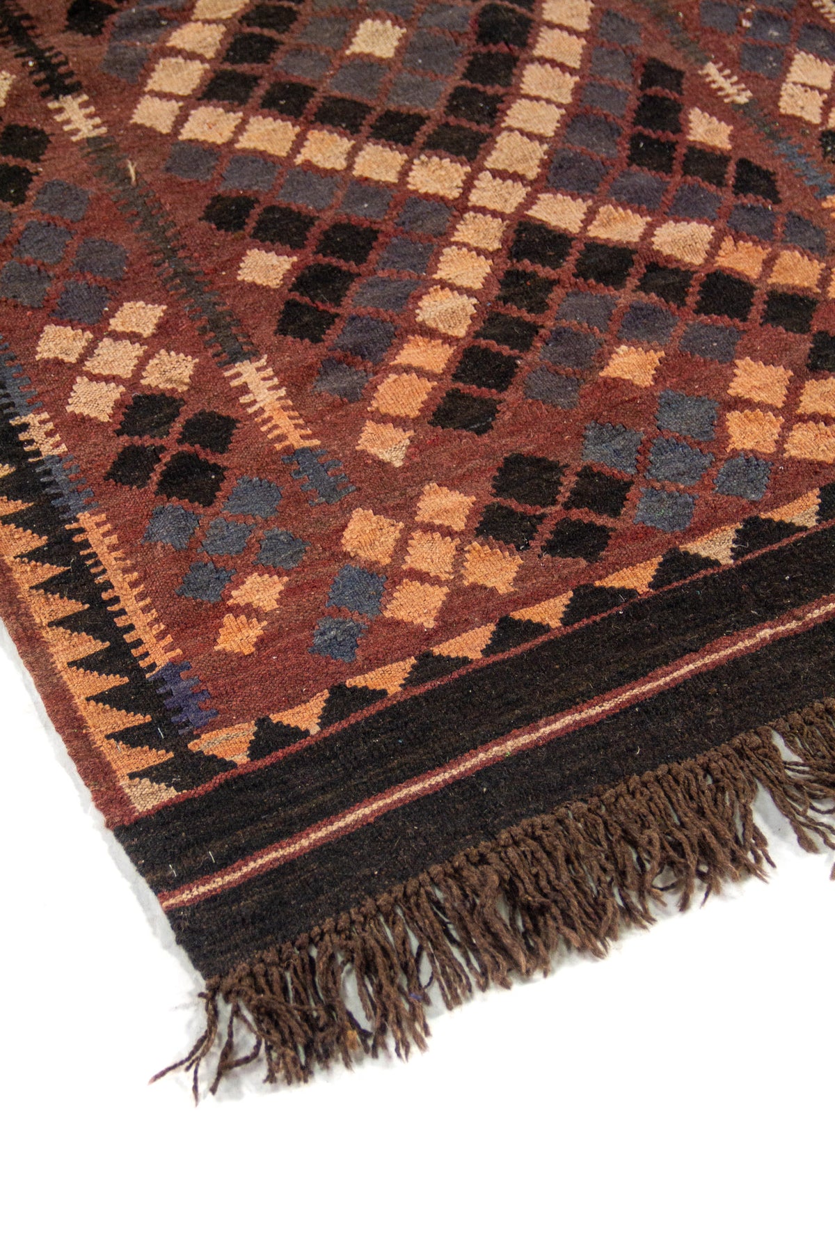 Detalle de esquina de alfombra artesanal con borde oscuro, motivos geométricos en rombos y flecos en tono marrón y beige.