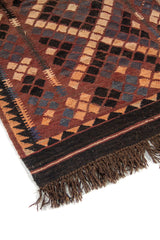 Detalle de esquina de alfombra artesanal con borde oscuro, motivos geométricos en rombos y flecos en tono marrón y beige.