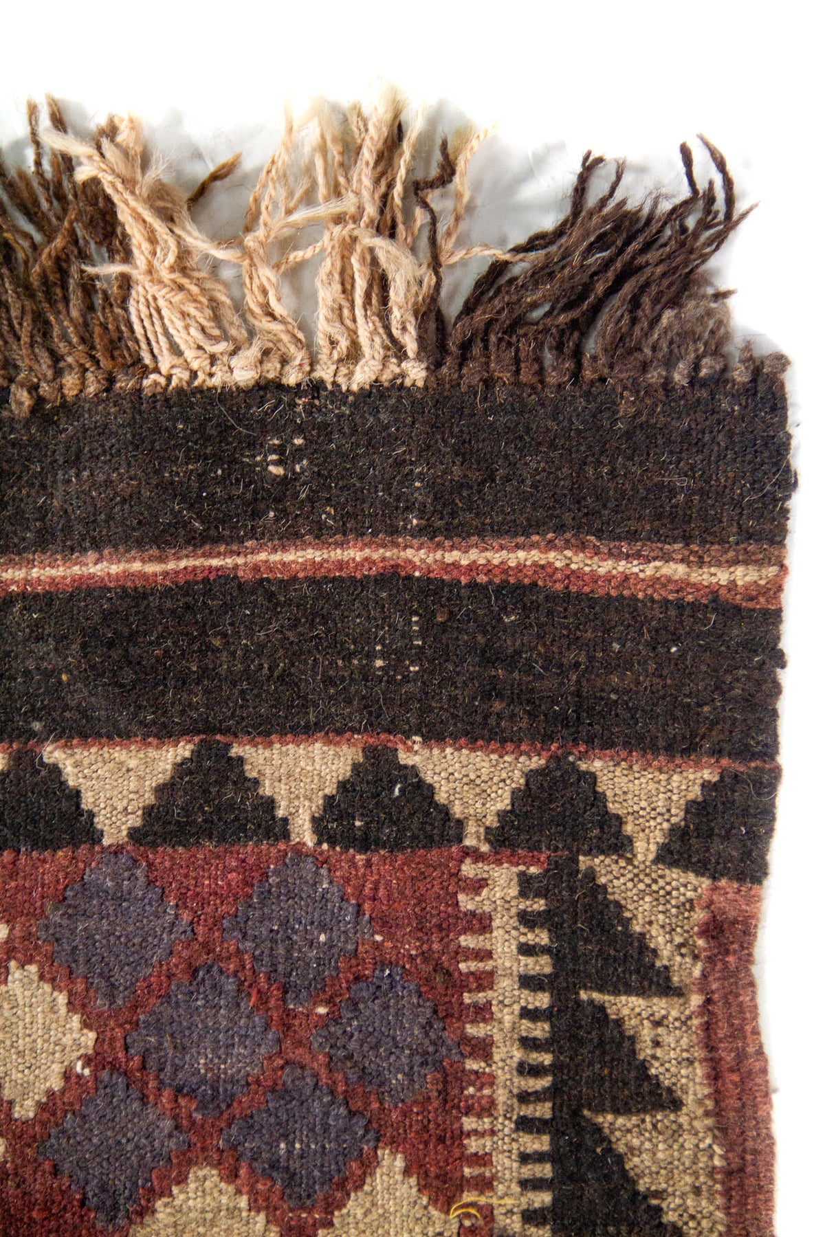 Detalle superior de alfombra tejida con flecos en tonos naturales y oscuros, y guarda geométrica en colores marrón, negro y beige.