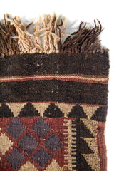 Detalle superior de alfombra tejida con flecos en tonos naturales y oscuros, y guarda geométrica en colores marrón, negro y beige.