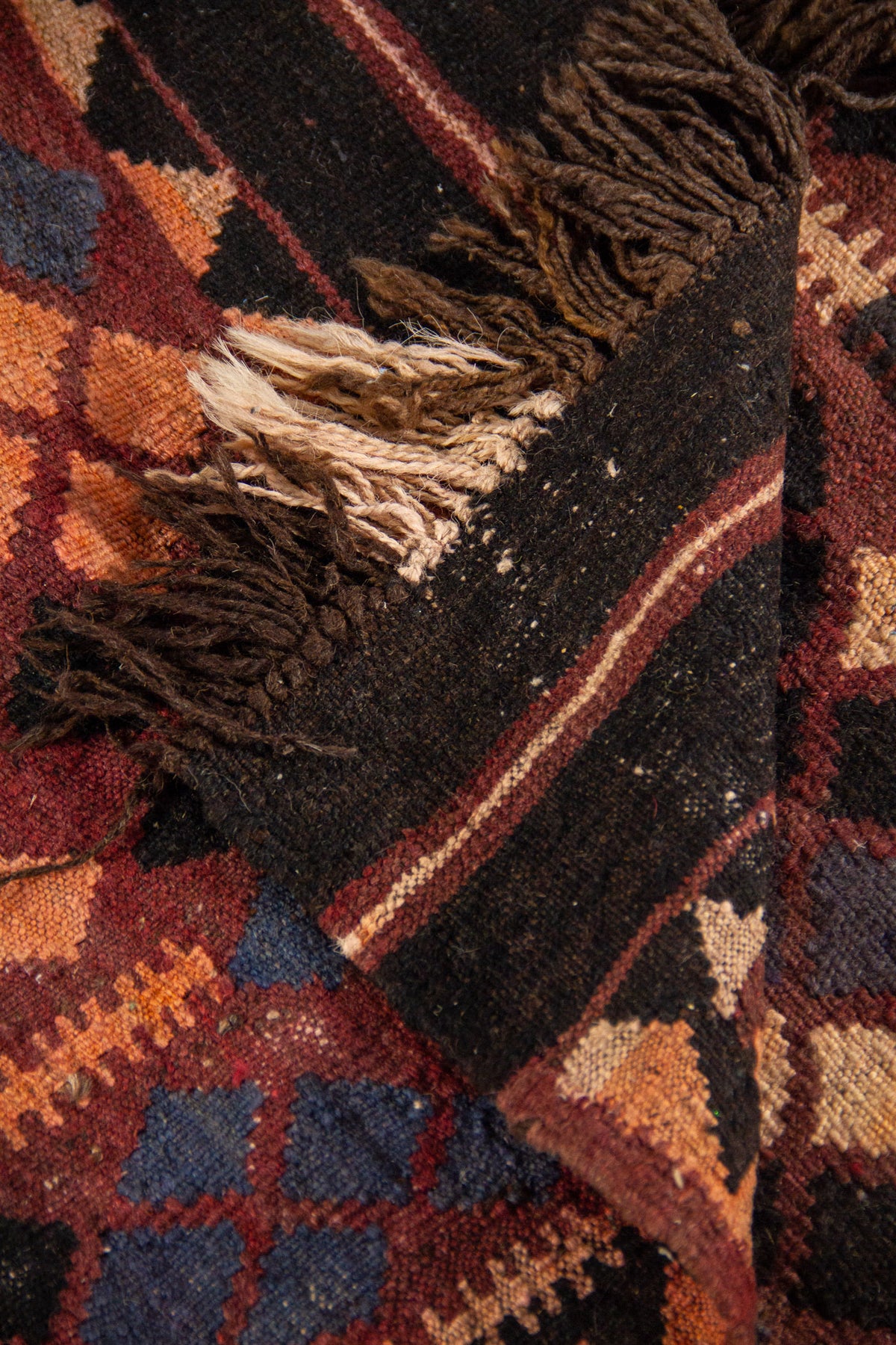 Detalle de alfombra doblada que muestra el reverso, los flecos y los patrones geométricos en tonos rojo, negro, beige y azul oscuro.